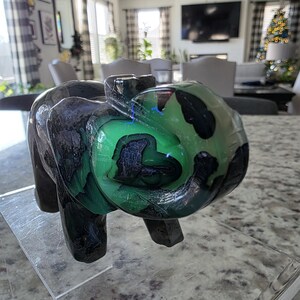 May include: A carved stone elephant figurine featuring a polished surface with deep black and vibrant green marbled patterns. The sculpture displays a smooth, glossy texture with natural color variations throughout the body and trunk. Text reads I. Michelle Crystals.