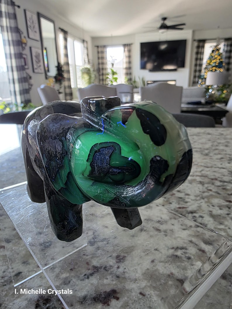 May include: A carved stone elephant figurine featuring a polished surface with deep black and vibrant green marbled patterns. The sculpture displays a smooth, glossy texture with natural color variations throughout the body and trunk. Text reads I. Michelle Crystals.