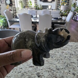 May include: A polished stone elephant figurine featuring a smooth, dark gray and black marbled surface with natural veining patterns. The carving depicts an elephant in a standing position with its trunk curved upward. Text reads: I. Michelle Crystals.