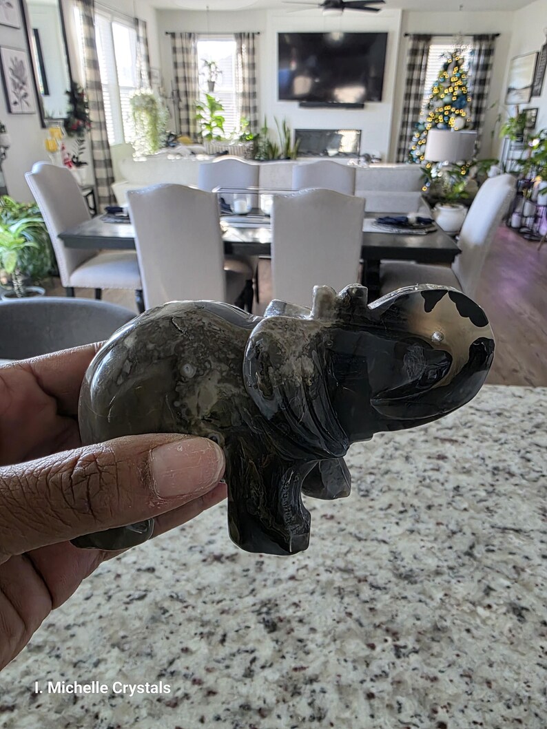 May include: A polished stone elephant figurine featuring a smooth, dark gray and black marbled surface with natural veining patterns. The carving depicts an elephant in a standing position with its trunk curved upward. Text reads: I. Michelle Crystals.