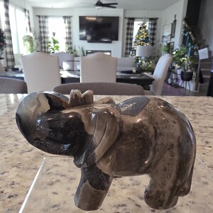 May include: A polished stone elephant figurine featuring swirling patterns of dark brown, gray, and cream colors. The sculpture is carved into a standing elephant shape with a raised trunk and is displayed on a clear acrylic stand with the text I. Michelle Crystals.