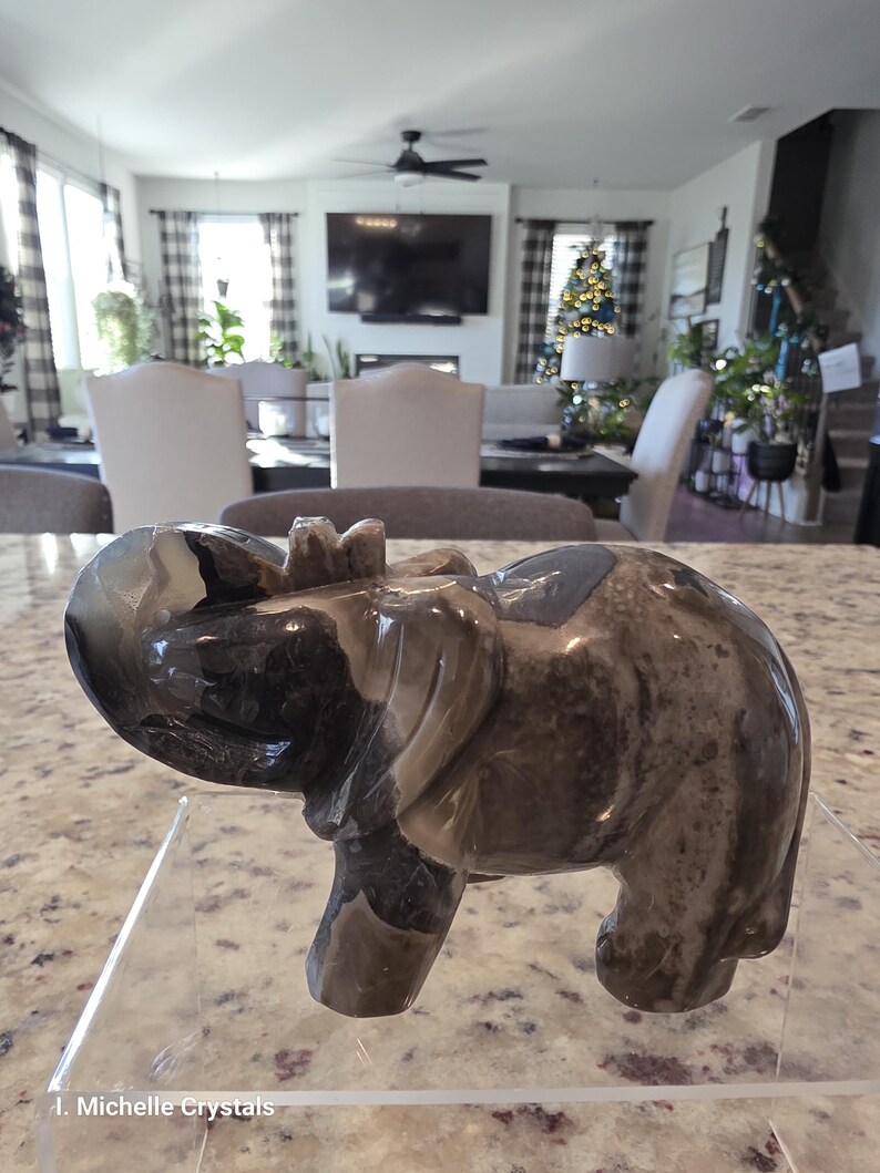 May include: A polished stone elephant figurine featuring swirling patterns of dark brown, gray, and cream colors. The sculpture is carved into a standing elephant shape with a raised trunk and is displayed on a clear acrylic stand with the text I. Michelle Crystals.