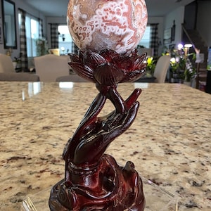 May include: A polished, spherical stone with intricate white and reddish-brown patterns resting atop a dark, reddish-brown carved wooden stand shaped like a lotus flower held by an upright hand. The text The Elevated Stone is visible at the base.