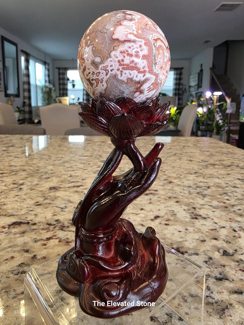 May include: A polished, spherical stone with intricate white and reddish-brown patterns resting atop a dark, reddish-brown carved wooden stand shaped like a lotus flower held by an upright hand. The text The Elevated Stone is visible at the base.