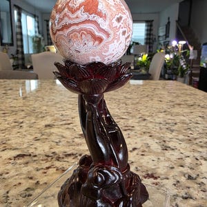 May include: A spherical stone with intricate swirling patterns in shades of cream, white, and reddish-brown, resting on a dark brown carved resin stand shaped like a lotus flower held by hands. The base features the text The Elevated Stone.