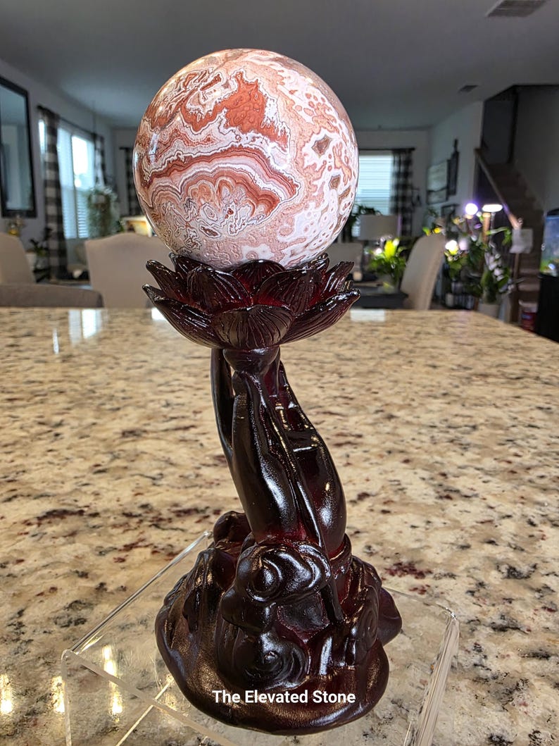 May include: A spherical stone with intricate swirling patterns in shades of cream, white, and reddish-brown, resting on a dark brown carved resin stand shaped like a lotus flower held by hands. The base features the text The Elevated Stone.