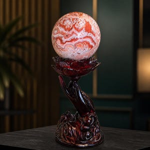 May include: A polished, spherical stone with intricate, swirling patterns of red, orange, and white. The sphere rests atop a dark, reddish-brown sculpture of an open hand holding a lotus flower, which serves as a decorative base for the stone orb.