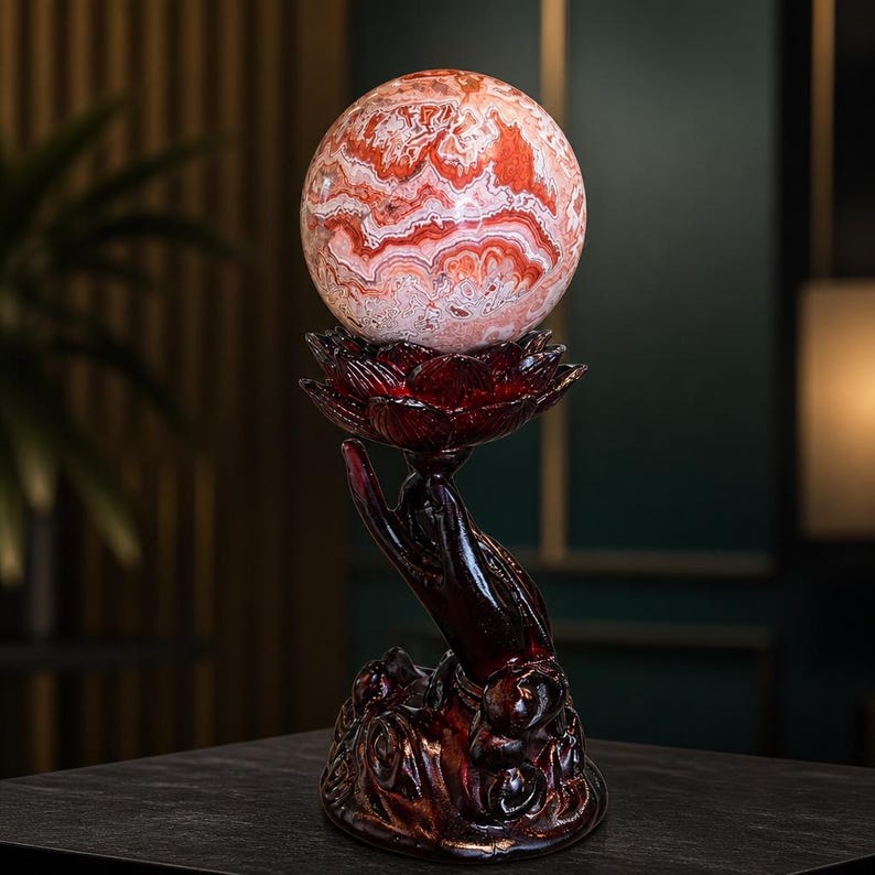May include: A polished, spherical stone with intricate, swirling patterns of red, orange, and white. The sphere rests atop a dark, reddish-brown sculpture of an open hand holding a lotus flower, which serves as a decorative base for the stone orb.