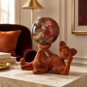 May include: A rust-colored, velvet-textured figurine of a reclining cat with its head resting on one paw. The cat figure holds a large, polished, spherical stone with swirling patterns of deep red, brown, and black colors above its body.