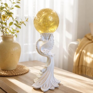May include: A white peacock-shaped sculpture serving as a base for a glowing, spherical crackle glass lamp. The bird figure features detailed plumage and an upright neck, supporting the illuminated yellow orb that emits a warm, soft light.