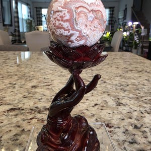 May include: A polished, spherical stone with swirling patterns of white, cream, and reddish-brown hues, resting atop a dark, glossy, reddish-brown stand shaped like a hand holding a lotus flower. The text "The Elevated Stone" is visible at the base.