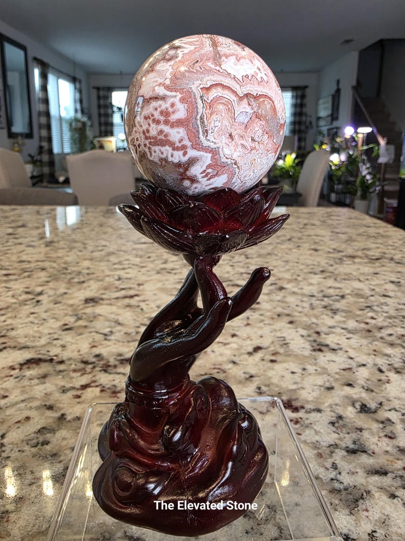 May include: A polished, spherical stone with swirling patterns of white, cream, and reddish-brown hues, resting atop a dark, glossy, reddish-brown stand shaped like a hand holding a lotus flower. The text "The Elevated Stone" is visible at the base.