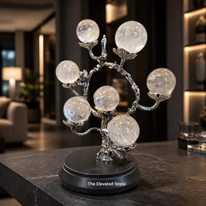 May include: A decorative sculpture featuring six clear, spherical crystal stones arranged on a silver-toned metal tree structure. The tree is mounted on a round black base with the text The Elevated Stone printed in white on the front surface.