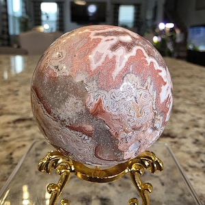 May include: A polished spherical stone featuring intricate patterns of reddish-brown, white, and grey mineral inclusions. The sphere rests on a decorative gold-colored metal tripod stand with curved legs. The text The Elevated Stone is visible.
