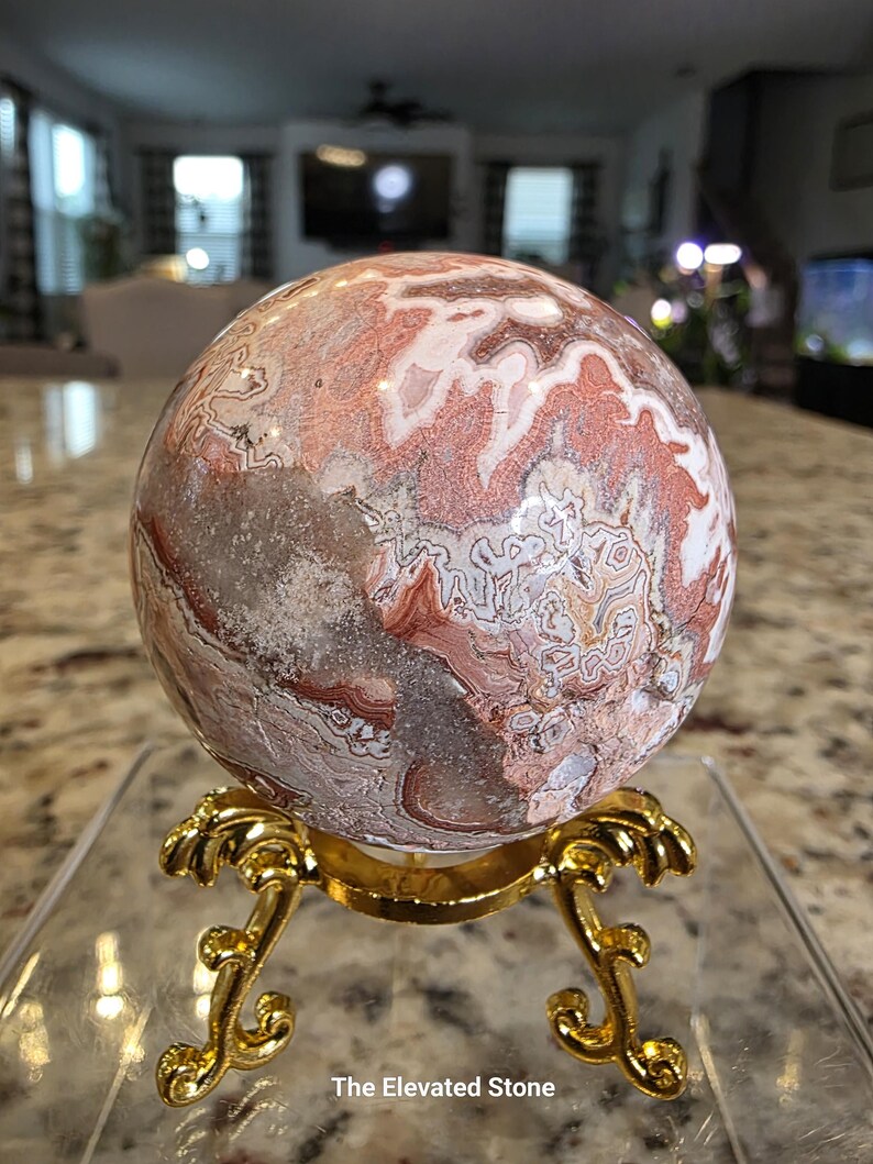 May include: A polished spherical stone featuring intricate patterns of reddish-brown, white, and grey mineral inclusions. The sphere rests on a decorative gold-colored metal tripod stand with curved legs. The text The Elevated Stone is visible.