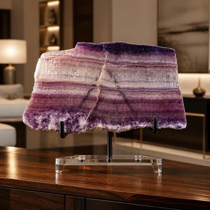 May include: A large, rectangular slab of banded fluorite mineral featuring horizontal layers of deep purple, lavender, and translucent white. The stone is mounted on a black metal stand with a clear acrylic base, displaying its natural crystalline texture.