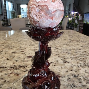 May include: A spherical stone with intricate pink, white, and brown swirling patterns resting on a dark reddish-brown stand shaped like a hand holding a lotus flower. The base features the text The Elevated Stone printed in white at the bottom.