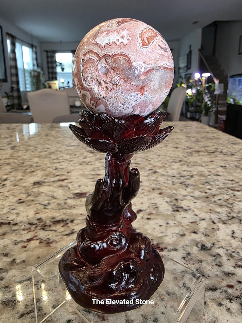 May include: A spherical stone with intricate pink, white, and brown swirling patterns resting on a dark reddish-brown stand shaped like a hand holding a lotus flower. The base features the text The Elevated Stone printed in white at the bottom.