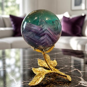 May include: A polished spherical fluorite gemstone featuring bands of deep purple, blue, and green hues. The sphere rests securely atop a gold-toned metal stand shaped like a delicate branch with textured leaves and a floral cup base.