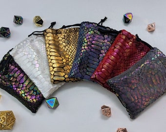 May include: A collection of seven drawstring dice bags in various colors, including white, gold, blue, red, and iridescent. The bags have a snakeskin pattern and are arranged in a semi-circle. Several polyhedral dice are scattered around the bags.