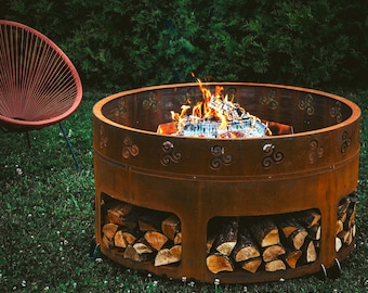 Può includere: Un focolare in metallo arrugginito con un fuoco di legna all'interno. Il focolare ha un design decorativo attorno alla parte superiore e un'area di stoccaggio per la legna da ardere sotto.