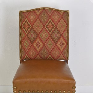 May include: A brown leather dining chair with a patterned fabric back. The chair has a carved wooden frame and brass nailhead trim.