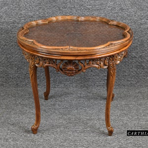 May include: A round, carved wooden table with a glass top. The table has a scalloped edge and intricate details. The table is on a gray background.