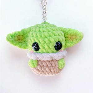 May include: A handmade green crocheted keychain featuring a character with large black eyes and large ears. The character is wearing a white and pink collar and is in a tan basket. The keychain is attached to a silver metal chain.