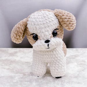 Puede incluir: Un perro de peluche hecho de hilo grueso y suave en tonos crema y marrón claro. El animal de peluche presenta grandes ojos redondos de plástico negro, una pequeña nariz negra bordada y una línea vertical para la boca, con orejas caídas.