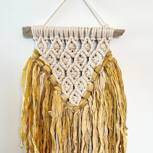 May include: A macrame wall hanging with a diamond pattern and a fringe of mustard yellow fabric.