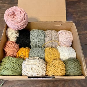 May include: An open cardboard box containing a selection of macrame cord in various colours. The colours include pink, black, green, yellow, and cream. The spools of cord are neatly arranged, ideal for crafting projects.