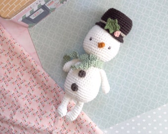 May include: A white crocheted snowman with a green scarf and a brown hat with a green star on top. The snowman is lying on a light blue and white patterned surface.