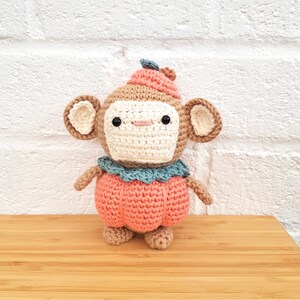 May include: A crocheted monkey wearing a peach-colored pumpkin costume with a blue flower on its hat. The monkey is standing on a wooden surface.