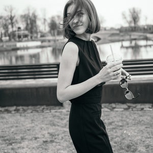May include: A woman in a black sleeveless dress holds a plastic cup with a straw and a beverage. She is standing outdoors in front of a body of water.