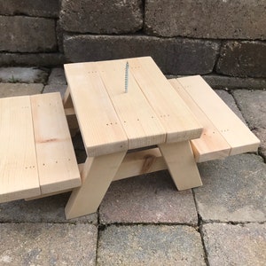 May include: A miniature wooden picnic table with two attached benches, constructed from light-colored natural wood planks. A single metal screw is vertically embedded in the center of the tabletop, which features visible nail heads on the surface.