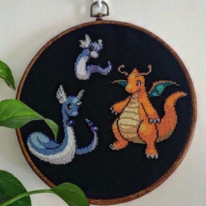 May include: Embroidered hoop art featuring three Pokemon characters: a blue and white Squirtle, a yellow and orange Charmander, and a blue and white Psyduck. The characters are stitched on a black background.