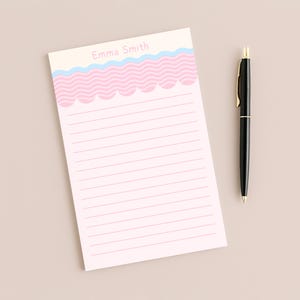 Peut inclure: Un bloc-notes rectangulaire avec des lignes horizontales roses et une bordure supérieure présentant une ligne ondulée bleu clair au-dessus d'un motif festonné rose. Le nom Emma Smith est imprimé en rose clair. Un stylo noir avec des détails dorés.