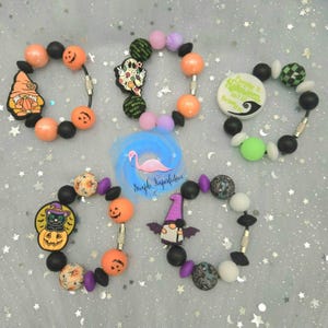 May include: Four Halloween themed bracelets with various charms, including a ghost, a pumpkin, a cat, and a gnome. The bracelets are made with black, orange, purple, green, and white beads.