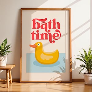Op de afbeelding: Een ingelijst print met de tekst "Bath time" in rode letters. Een gele rubberen eend drijft in een blauw bad.