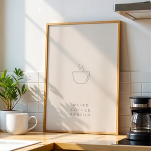 May include: A framed print with a minimalist design of a coffee cup and the text "Weird Coffee Person" on a light beige background.