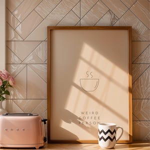 May include: A framed print with a minimalist design of a coffee cup with steam rising from it. The text "Weird Coffee Person" is below the cup. The print is hanging on a wall with beige and white tiles.