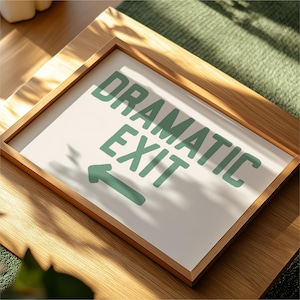 Op de afbeelding: Een ingelijst bord met de woorden "DRAMATIC EXIT" in groene blokletters en een pijl die naar links wijst. Het bord heeft een witte achtergrond en een lichtbruine houten lijst. Het bord staat op een houten oppervlak.