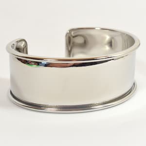 Può includere: Un braccialetto rigido in argento con una finitura liscia e lucida. Il braccialetto è aperto e ha un design semplice e moderno.