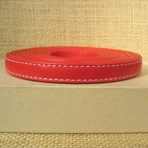 May include: A roll of red leather cord with white stitching. The cord is rolled up and is sitting on a brown surface.