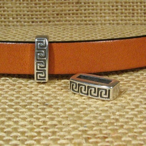 May include: Two silver metal belt buckle keepers with a Greek key design. The keepers are on a brown leather strap.