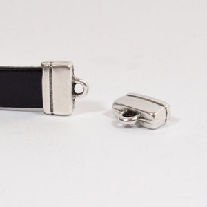 May include: Silver-toned metal clasp for leather straps. The clasp has a rectangular shape with a small loop on the top. The clasp is designed to be used with leather straps.