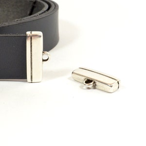 May include: Two silver metal belt buckle closures. One closure is attached to a black leather belt. The closures have a rectangular shape with a small loop on the top.