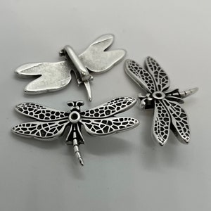 May include: Three silver dragonfly charms with intricate details. The charms are in various positions, showcasing their delicate wings and body.