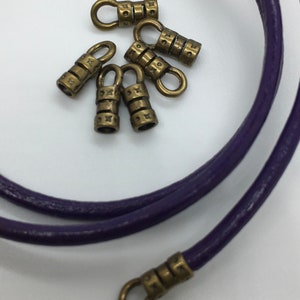 May include: A close-up of seven antique gold-toned metal cord ends with a decorative design. The cord ends have a small loop at the top and are designed to be used with leather cord.