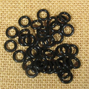 Könnte beinhalten: Eine Ansammlung kleiner, schwarzer Gummi-O-Ringe auf einer beigen, strukturierten Oberfläche. Die O-Ringe sind kreisförmig mit einem hohlen Zentrum und scheinen aus einem flexiblen Material zu bestehen. Die Beleuchtung hebt die glatte Textur der Ringe hervor.